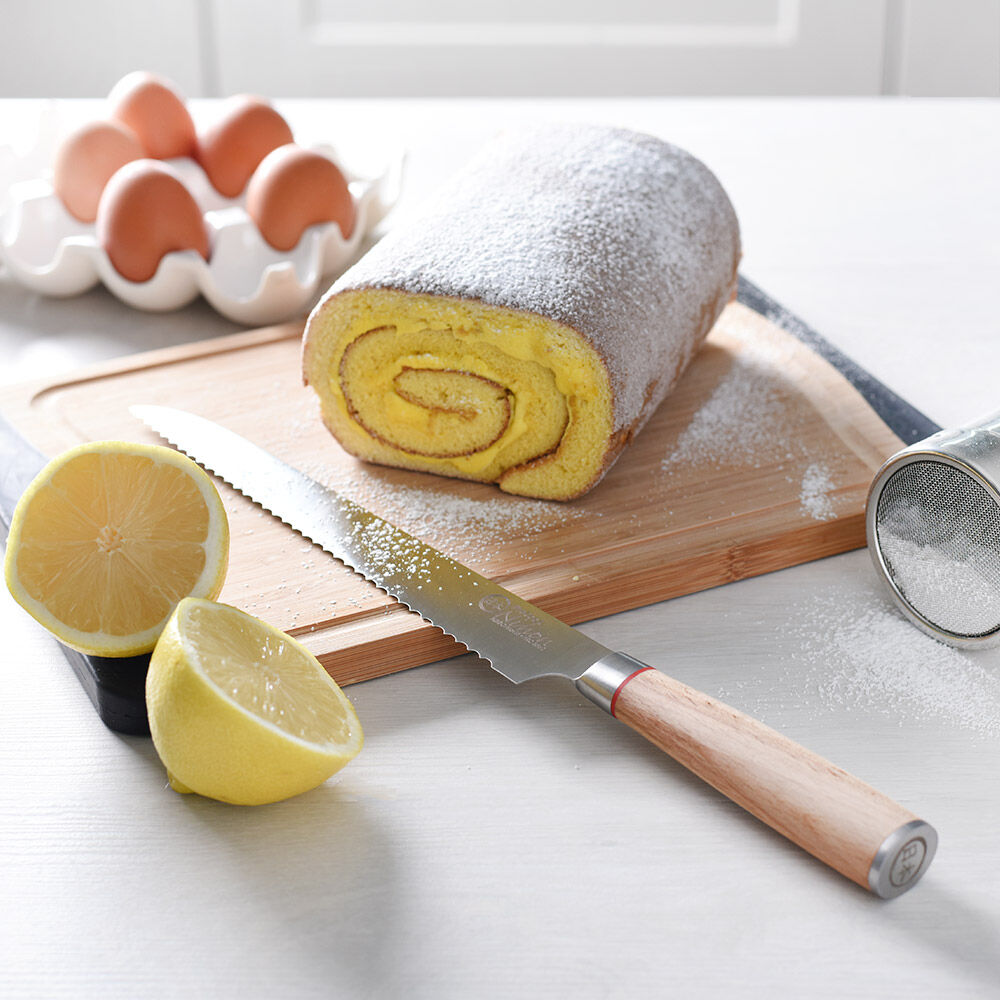 Image of ProCook Nihon X50 bread knife resting on a chopping board next to a jam roly poly desert